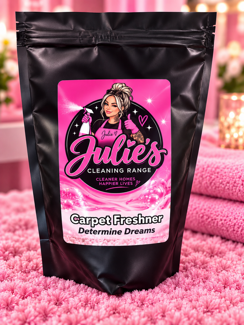 A stand-up resealable pouch of Julie's Carpet Freshner from the Cleaning Range, featuring a black bag with a bright pink label, set on a pink shag rug with folded pink towels in the background.